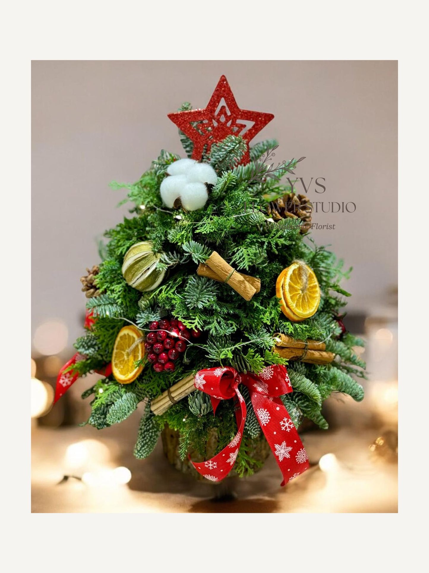 Rustic Christmas table top tree with pinecones and ribbons on a wooden table, with 'YVS Florist Studio' in the background.