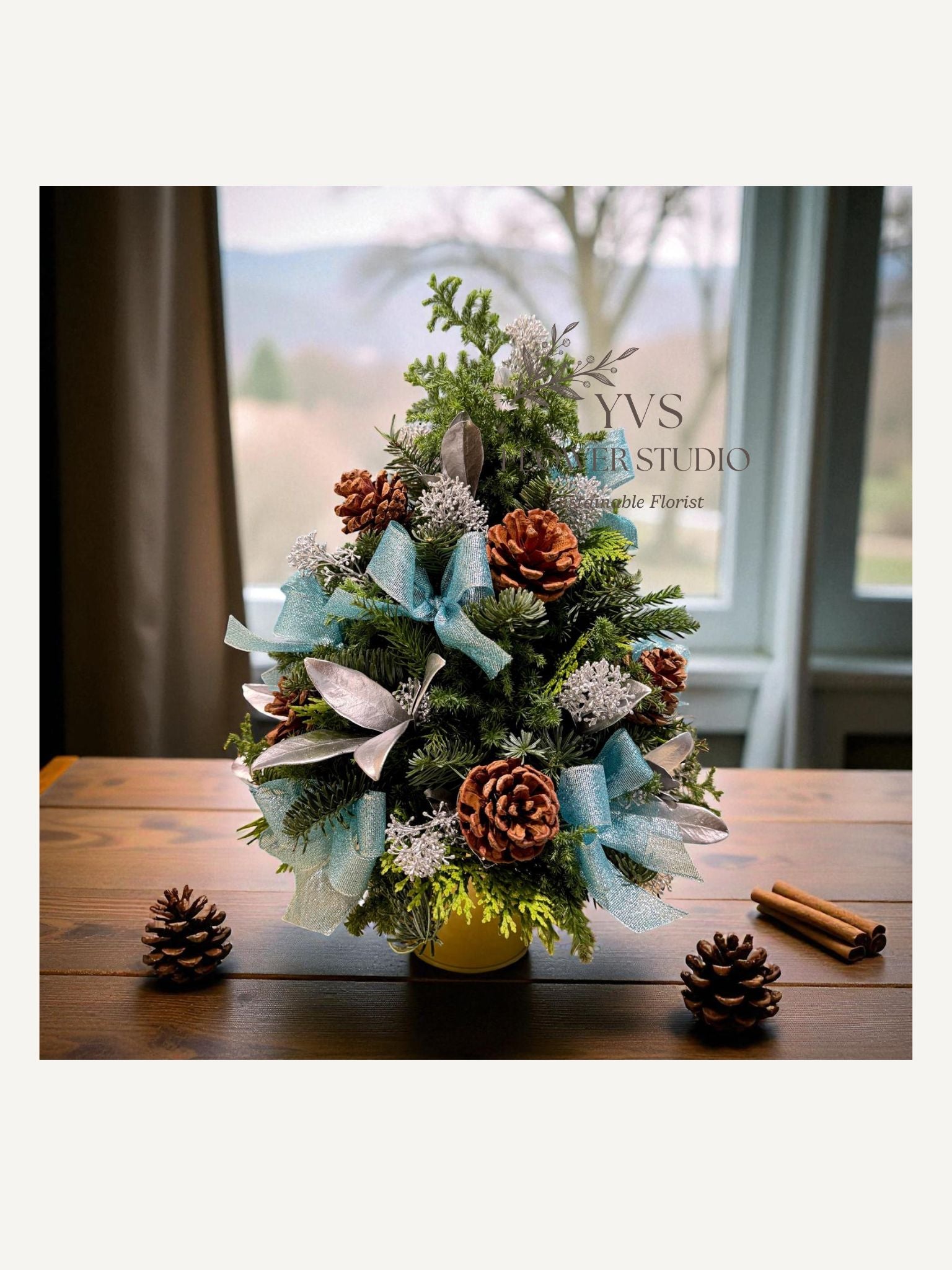 Blue and silver Christmas table top tree with pinecones and ribbons on a wooden table, with 'YVS Florist Studio' in the background.